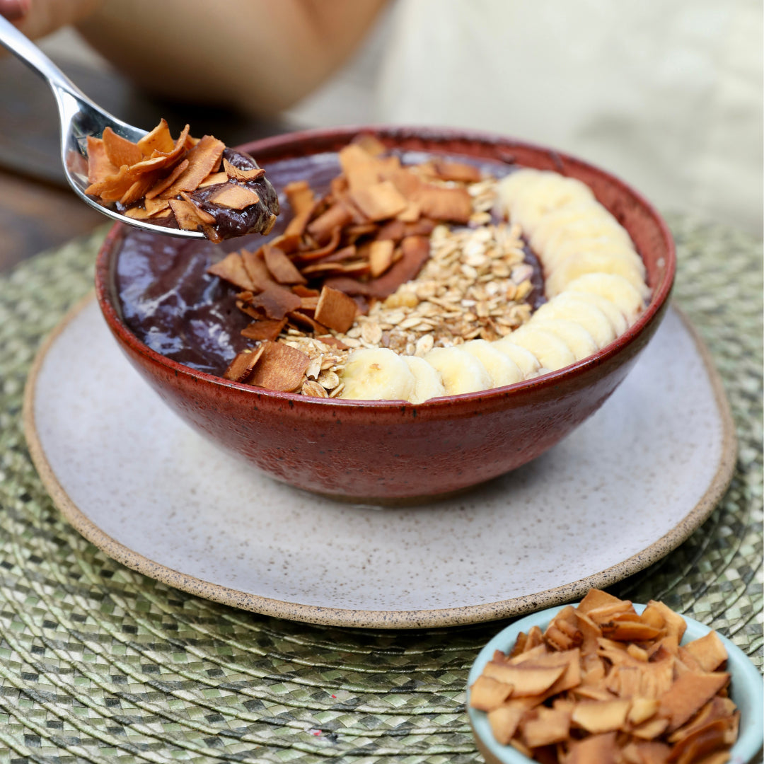 Tigela de açaí com banana em rodelas, granola e chips de coco crocante sabor cacau por cima.