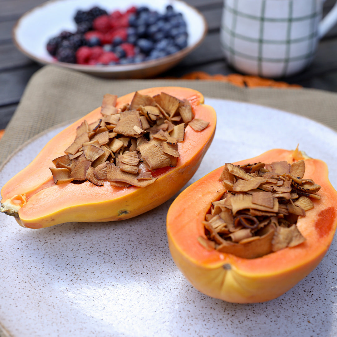 Duas fatias de mamão papaya, com chips de coco crocante em cima, e um prato com mertilo, amora e framboesa de fundo.